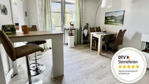 A modern, well-lit room with a dining area, a small table with a decorative item, and a window with curtains. - Waldläufer Brilon (Brilon)