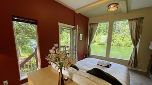 A bedroom with a bed, a wooden dresser, a vase with flowers, and a view of a garden through large windows.