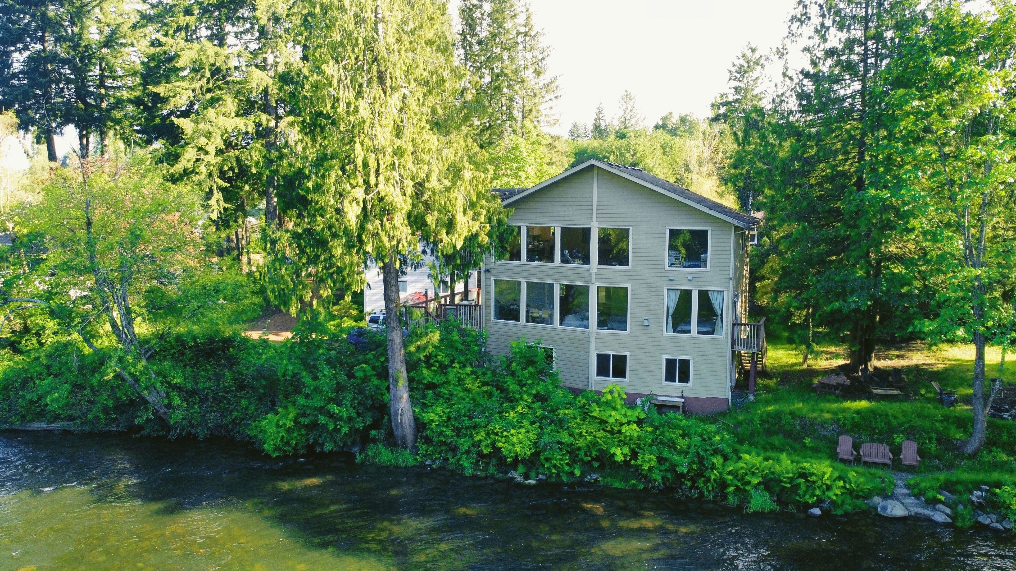 A two-story house with a large window on the second floor, surrounded by dense greenery and a river.