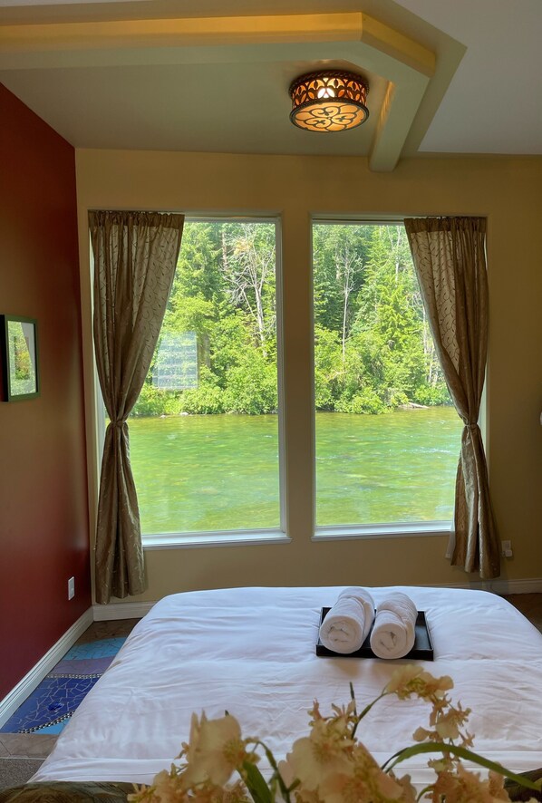 Habitación de hotel con una cama, dos ventanas con cortinas, una lámpara de techo y vista al río.