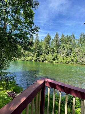 Ein Fluss mit einer Holzterrasse und Sitzgelegenheiten, umgeben von dichtem Grün und einem klaren Himmel.