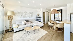 Uma sala de estar moderna com um sofá branco, uma área de jantar com cadeiras de vime e uma cozinha visível através de uma porta aberta.