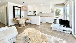 Uma sala de estar moderna com sofá branco, área de refeições com cadeiras de vime, cozinha com armários brancos e uma grande janela com vista para o exterior.