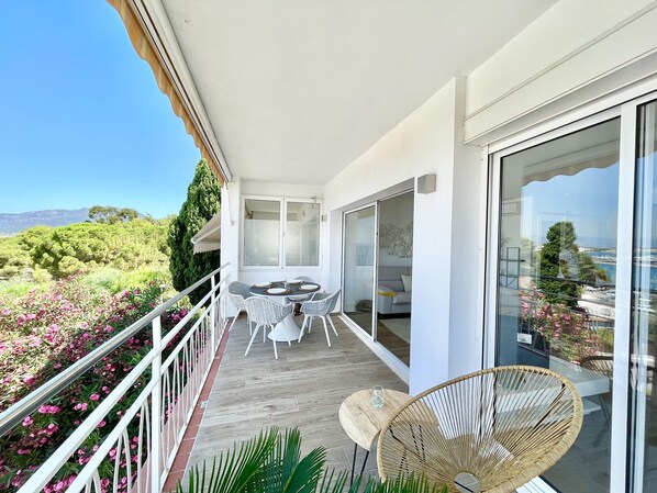 Een balkon met een tafel en stoelen, een rieten stoel en uitzicht op zee en bergen.