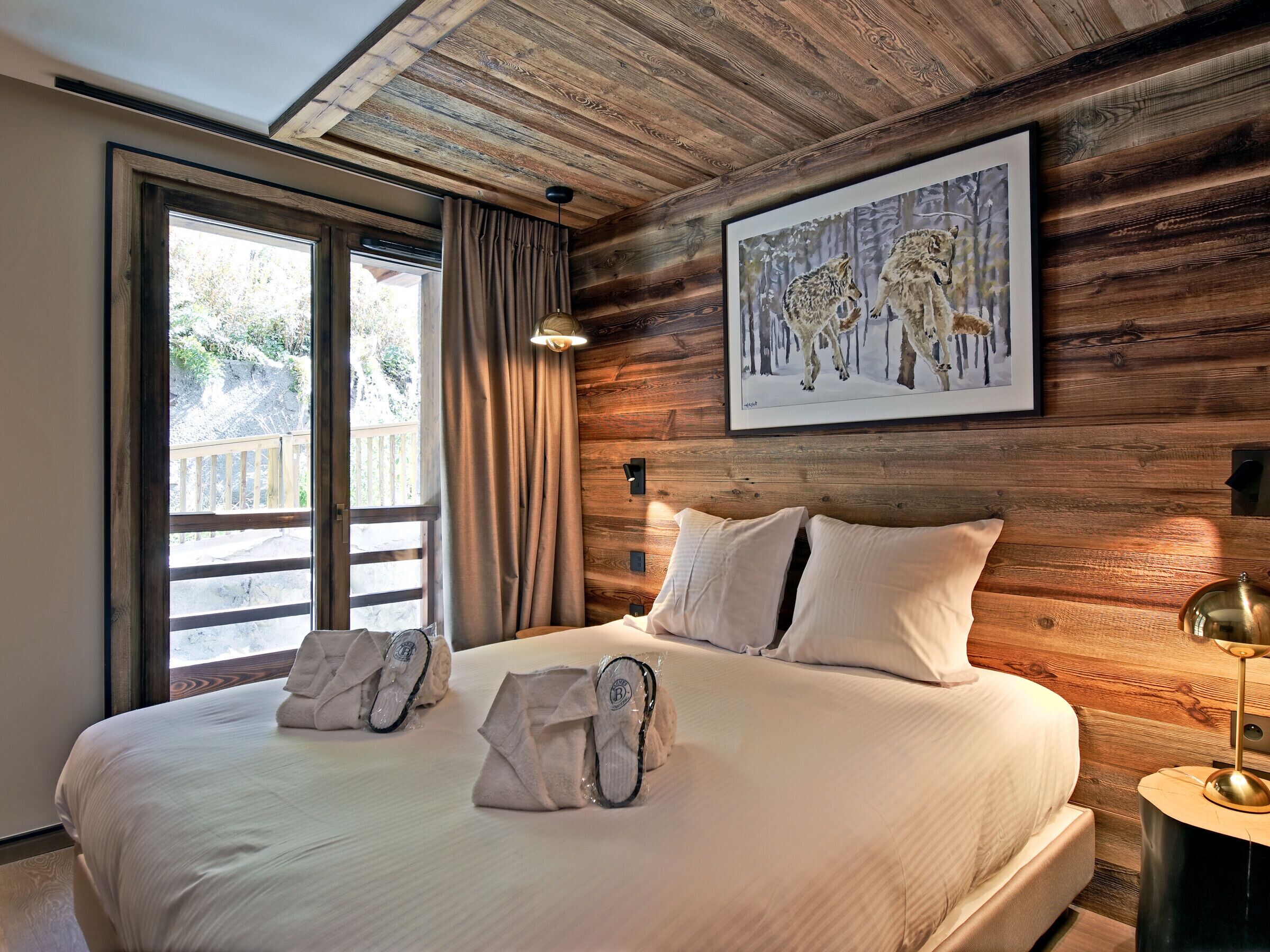 A hotel room with a wooden wall, a large bed with white linens, a painting of wolves, and a view through a window.