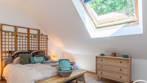 A bedroom with a sloped ceiling, a skylight, a bed with patterned bedding, a bedside table, a wicker chair, a dresser, and a small rug. - Cozy Gîte with Shared Heated Pool near Angers (Tiercé)