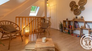 A cozy loft bedroom with a skylight, wooden flooring, wicker chairs, a small table, and a bookshelf with books and decorative items. - Cozy Gîte with Shared Heated Pool near Angers (Tiercé)