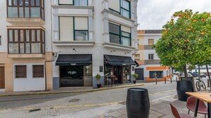 A street view with a building featuring a storefront with a sign that reads "Vinos." There are outdoor seating areas with tables and chairs, and a tree with orange fruits. - El Rincón del Baluarte by H - Centro Ayamonte AVHVIIISP (Ayamonte)
