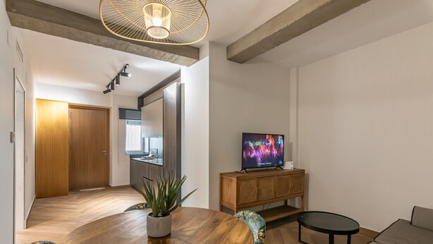 A modern living room with a wooden dining table, a sofa, a TV on a wooden cabinet, and a kitchen in the background. - El Rincón del Baluarte by H - Centro Ayamonte AVHVIIISP (Ayamonte)