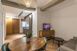 A modern living room with a wooden dining table, a sofa, a TV on a wooden cabinet, and a kitchen in the background.