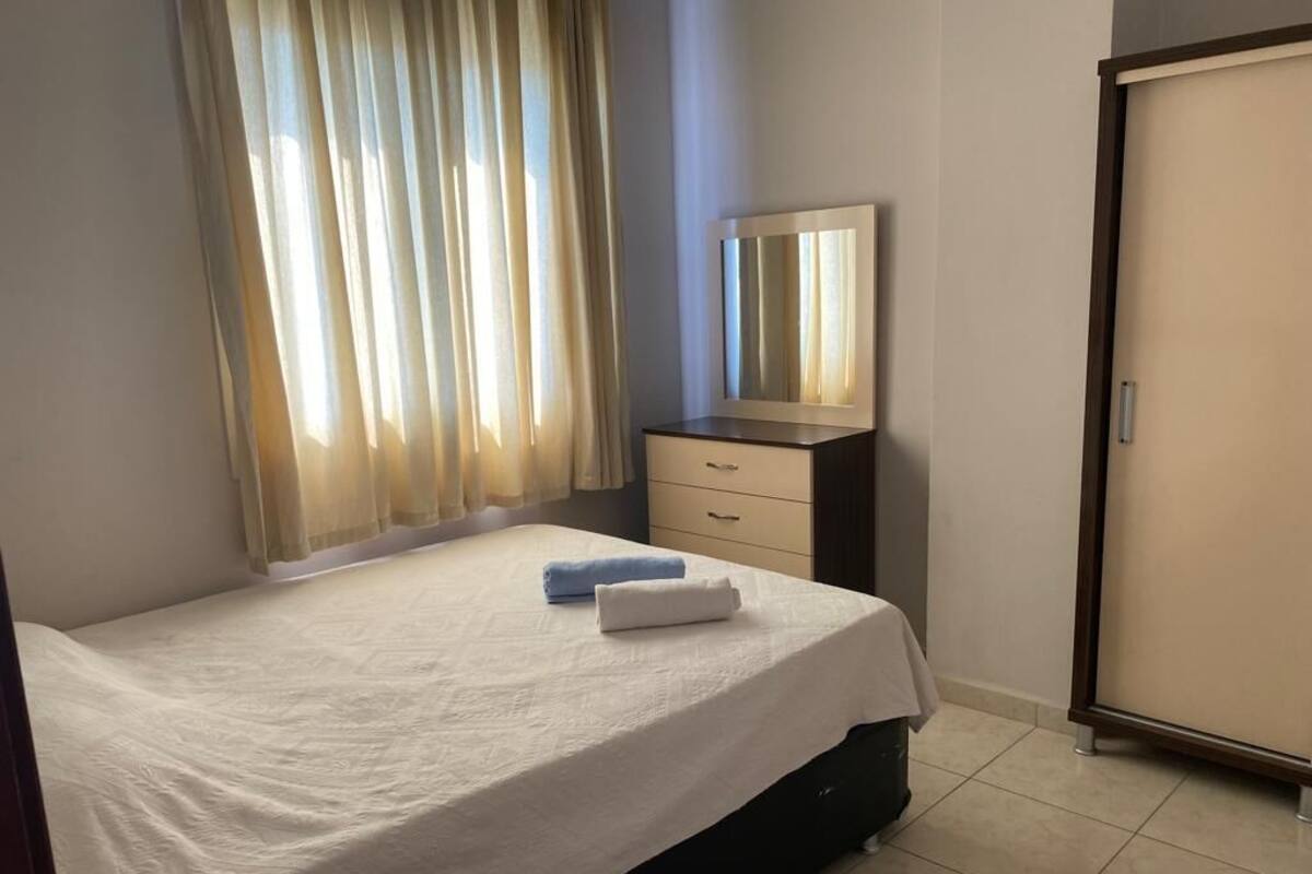Ein Schlafzimmer mit Bett, Nachttisch mit Spiegel, Kleiderschrank und Fenster mit Vorhängen.