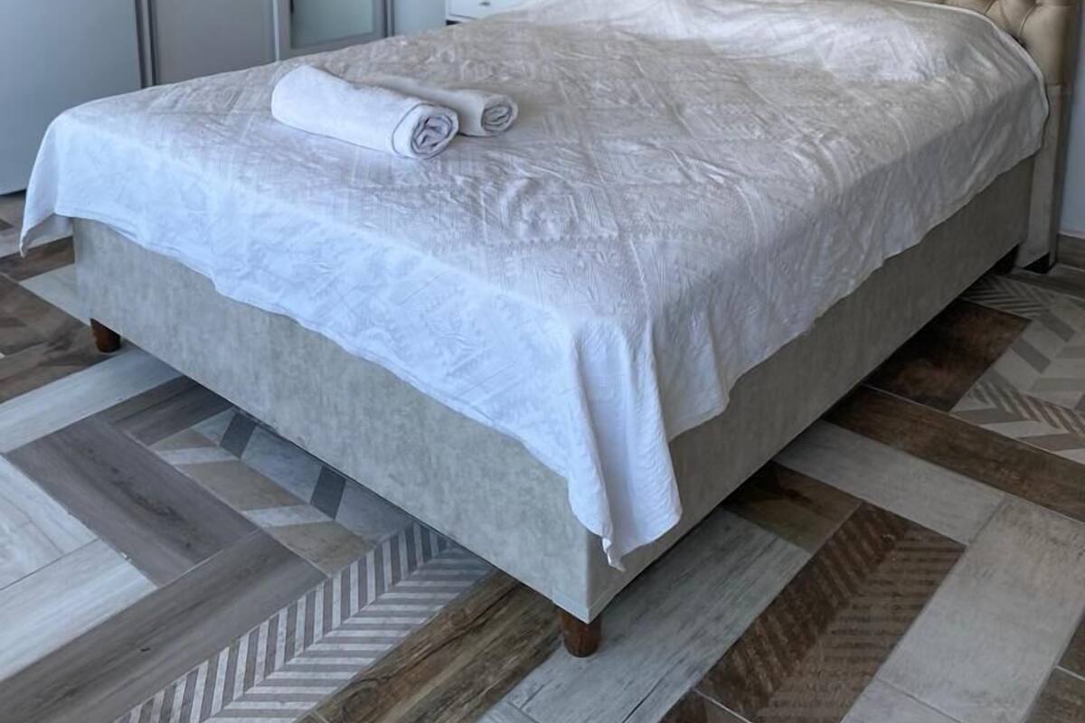 Ein Schlafzimmer mit einer gepolsterten Kopfteile, einem ordentlich bezogenen Bett mit einem aufgerollten weißen Handtuch, einem weißen Nachttisch und einem Kleiderschrank.