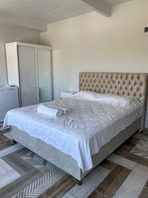 A neatly made bed with a white bedspread and two rolled towels, a tufted headboard, and a herringbone-patterned floor.