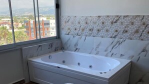 Ein Badezimmer mit einer großen Badewanne, einem Fenster mit Blick auf die Stadt und Fliesen mit Musterung an der Wand.
