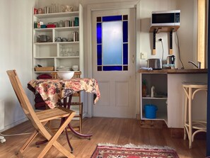 Une cuisine compacte équipée d’un four à micro-ondes, d’une bouilloire et d’un petit évier. On aperçoit un coin repas avec une chaise et une table, ainsi qu’une bibliothèque remplie de divers objets.