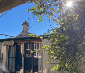 Une petite maison avec une cheminée, une fenêtre ornée d’un rideau et une branche d’arbre au premier plan.