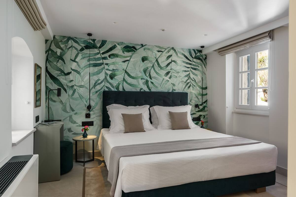 A bedroom with a large bed, a headboard, a small bedside table, a lamp, and a window.