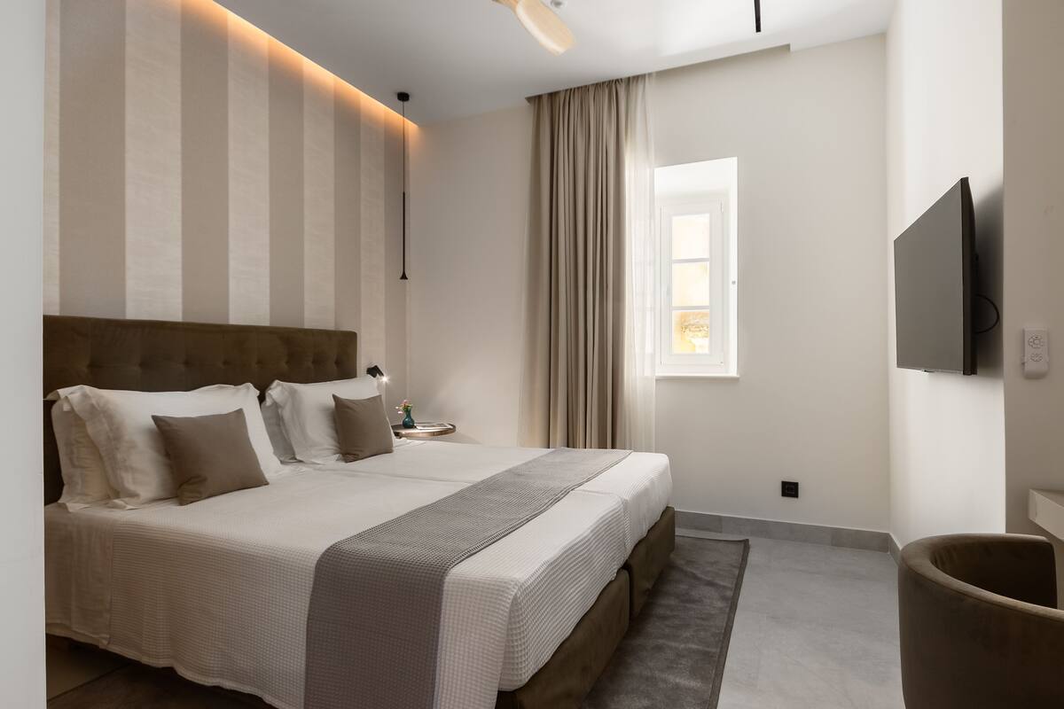 A neatly made bed with a headboard, a bedside table with a lamp, a television mounted on the wall, and a window with curtains.