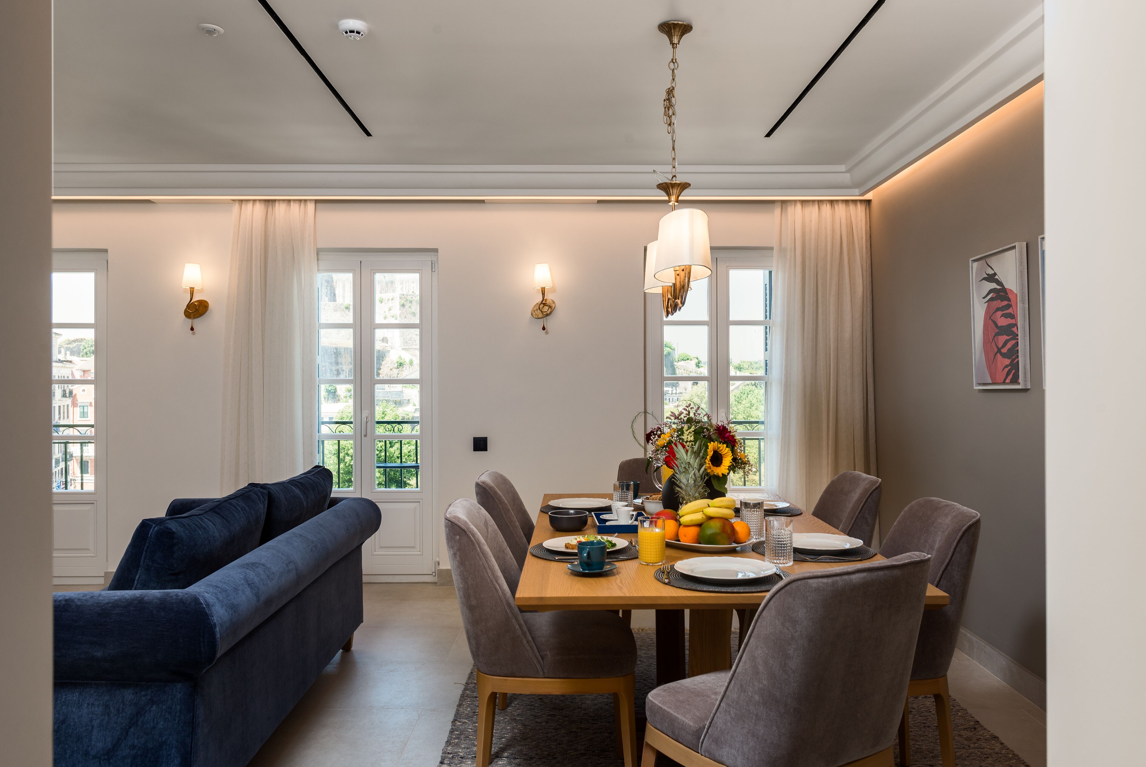 A dining area with a wooden table set for four, surrounded by chairs, with a sofa and curtains in the background.