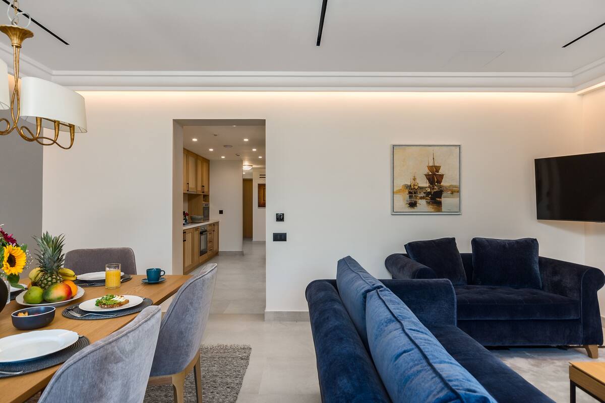 A modern living room with a dark blue sofa, a dining table with plates and a fruit bowl, and a kitchen in the background.