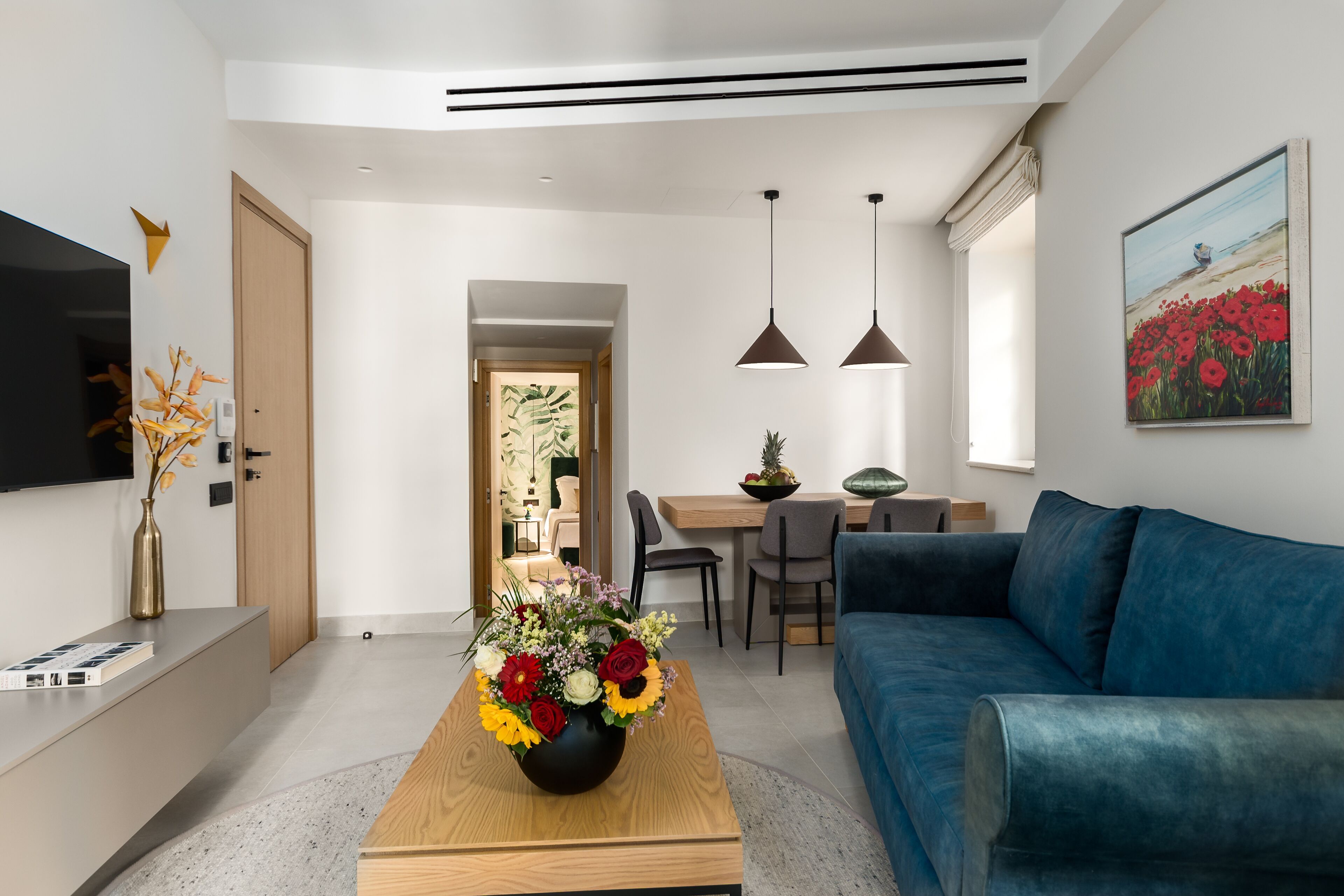 Una sala moderna con un sofá azul, una mesa de centro de madera con un jarrón floral, un comedor con una mesa y sillas de madera, y un cuadro en la pared.