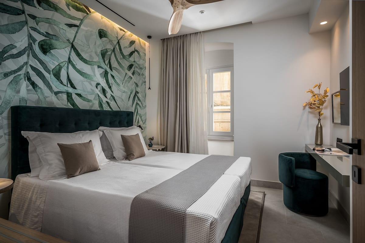 A modern bedroom with a large bed, a headboard with leaf patterns, a bedside table, a nightstand, a desk, a chair, and a window with curtains.