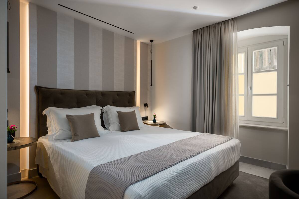 A neatly made bed with a headboard, a bedside table with a lamp, and a window with curtains.
