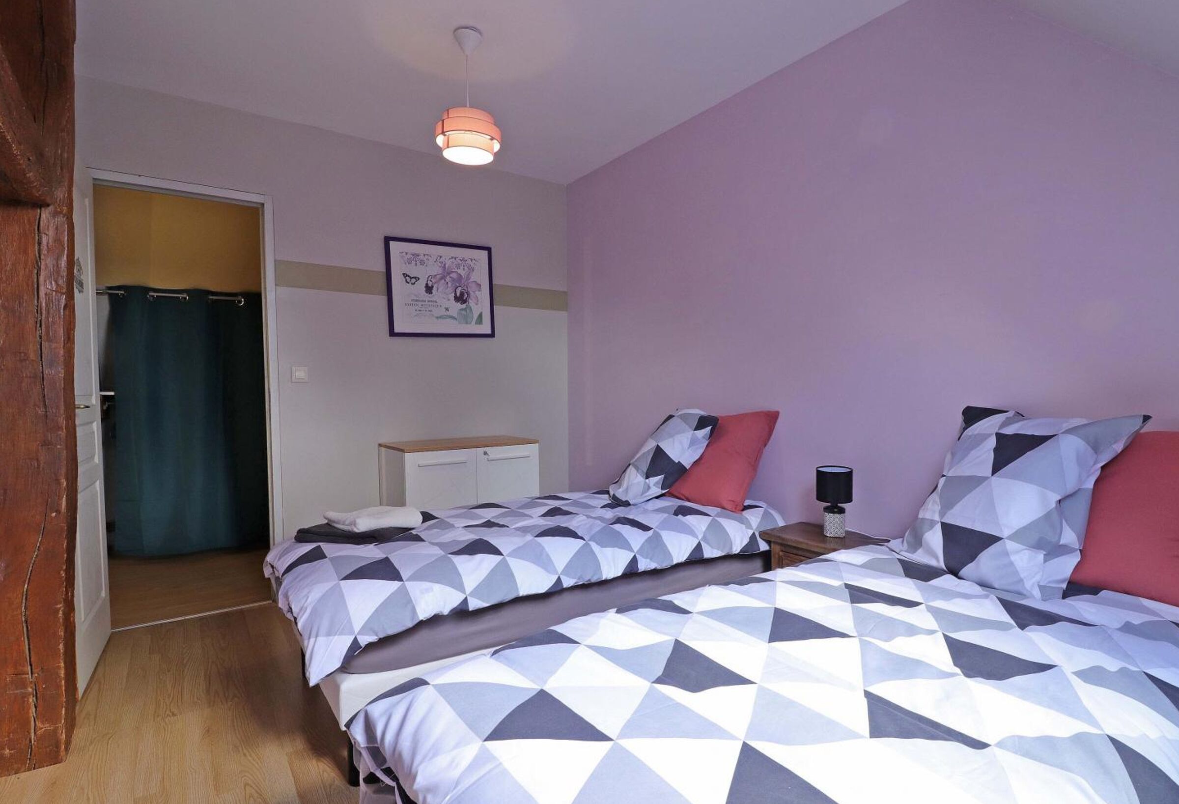 A bedroom with two beds, a nightstand, a lamp, a framed picture on the wall, and a door leading to another room.