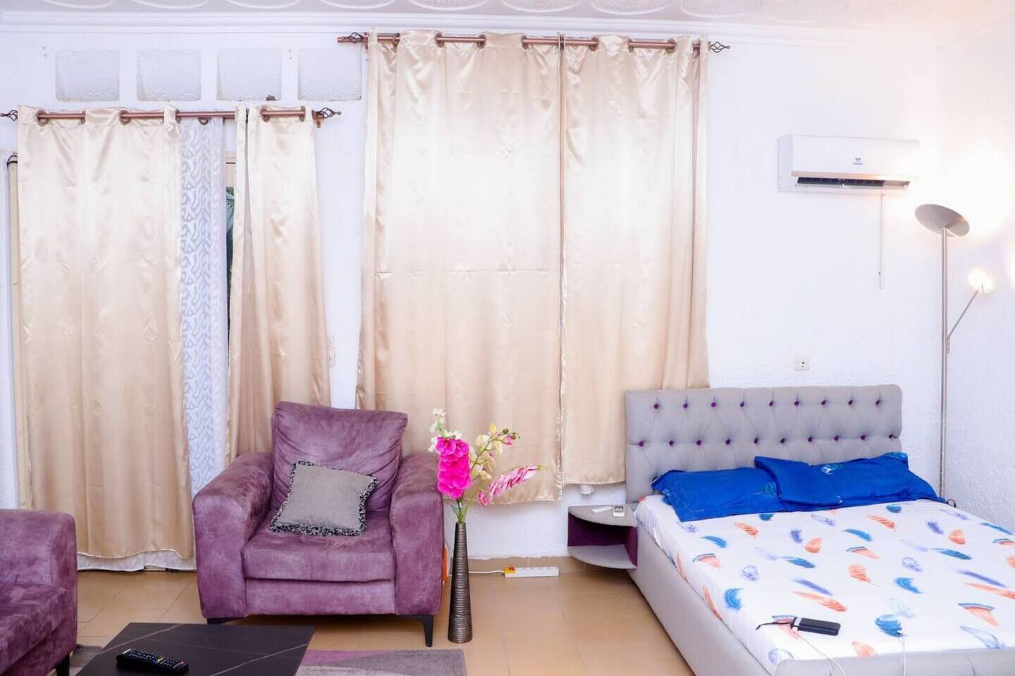 Une chambre à coucher comprenant un lit, une chaise, une petite table, un vase avec des fleurs et un appareil de climatisation.