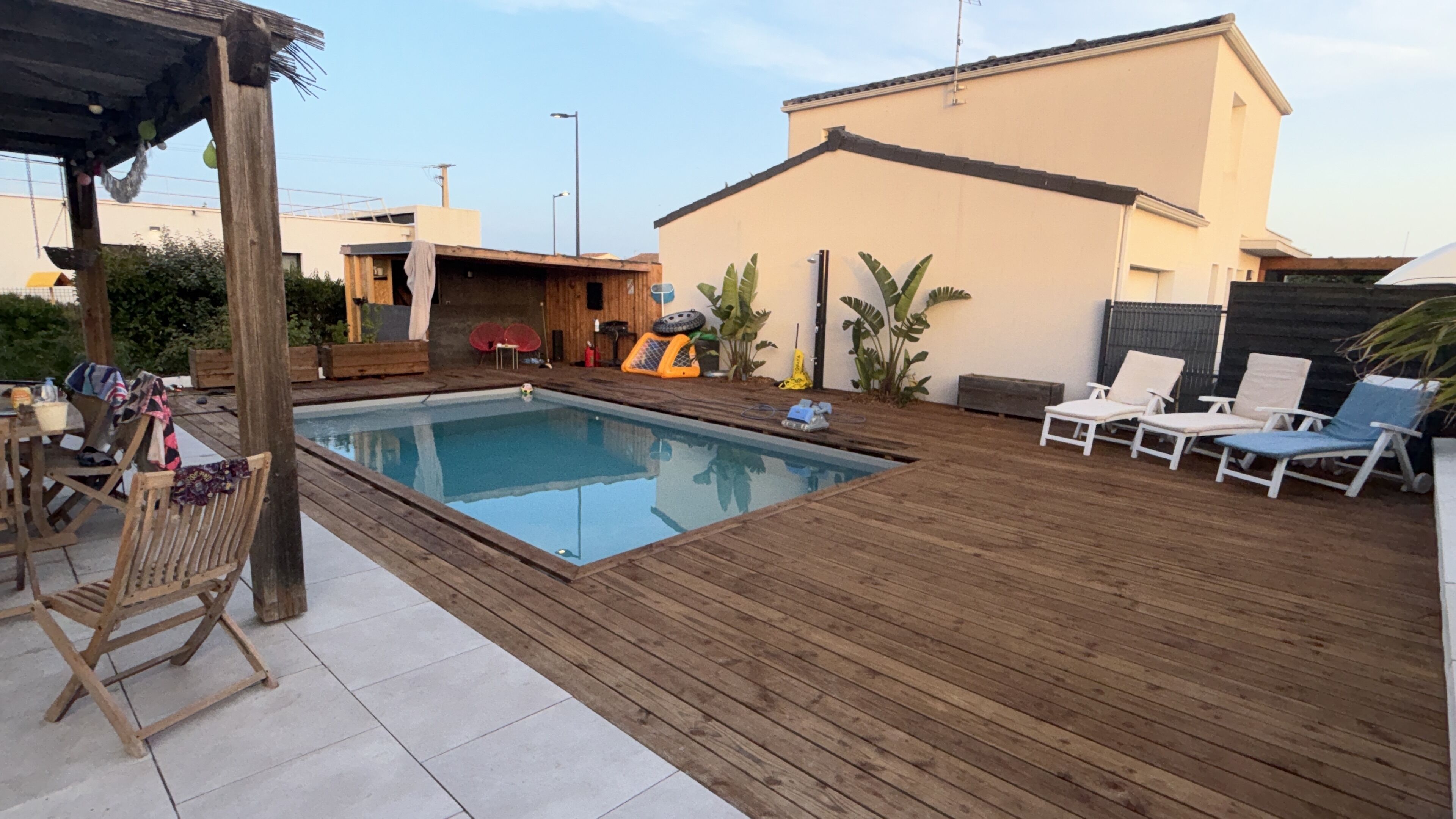 Ein Schwimmbad mit Holzterrasse, Liegestühlen und einem überdachten Bereich mit Tisch und Stühlen.