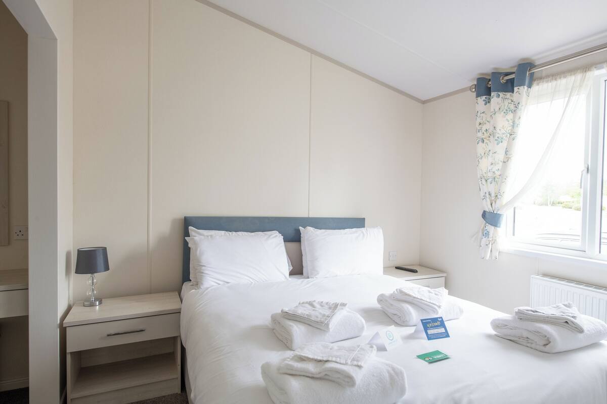 Una habitación de hotel con cama, mesita de noche, lámpara y ventana con cortinas.