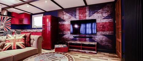 Een woonkamer met een rode bank, een ronde salontafel met een Union Jack dessin, een flatscreen-televisie bevestigd aan een muur met een bakstenen patroon, en een rode koelkast.
