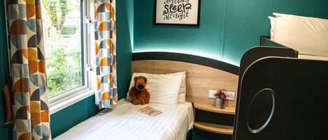 A bedroom with a bed, a small bedside table, a teddy bear, a framed quote on the wall, and a window with curtains.