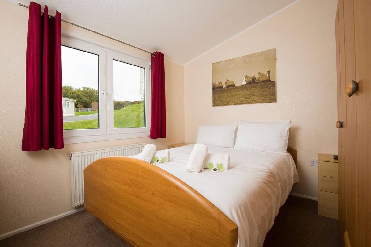 Un dormitorio con cama, ventana con cortinas, radiador, un cuadro en la pared y una puerta de madera.