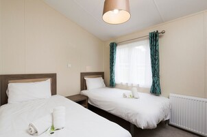 Habitación de hotel con dos camas individuales, cabecero de madera, mesita de noche, lámpara y ventana con cortinas.