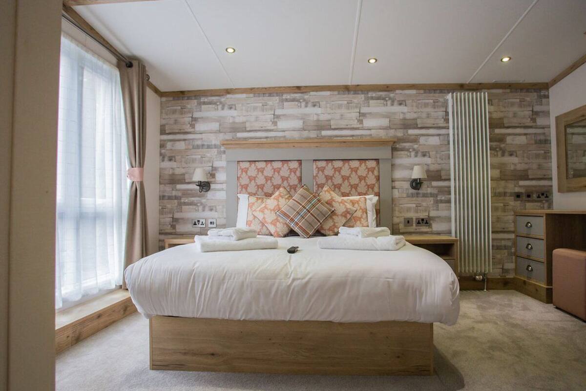 Un dormitorio con una cama grande, un cabecero de madera, una pared con diseño de piedra, una ventana con cortinas, una mesita de noche con una lámpara y un aparador.
