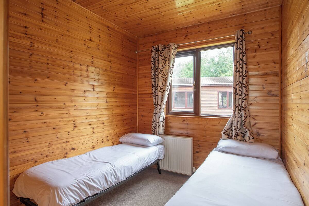 Una habitación de cabaña pequeña con dos camas individuales, paredes de madera, una ventana con cortinas y un radiador.