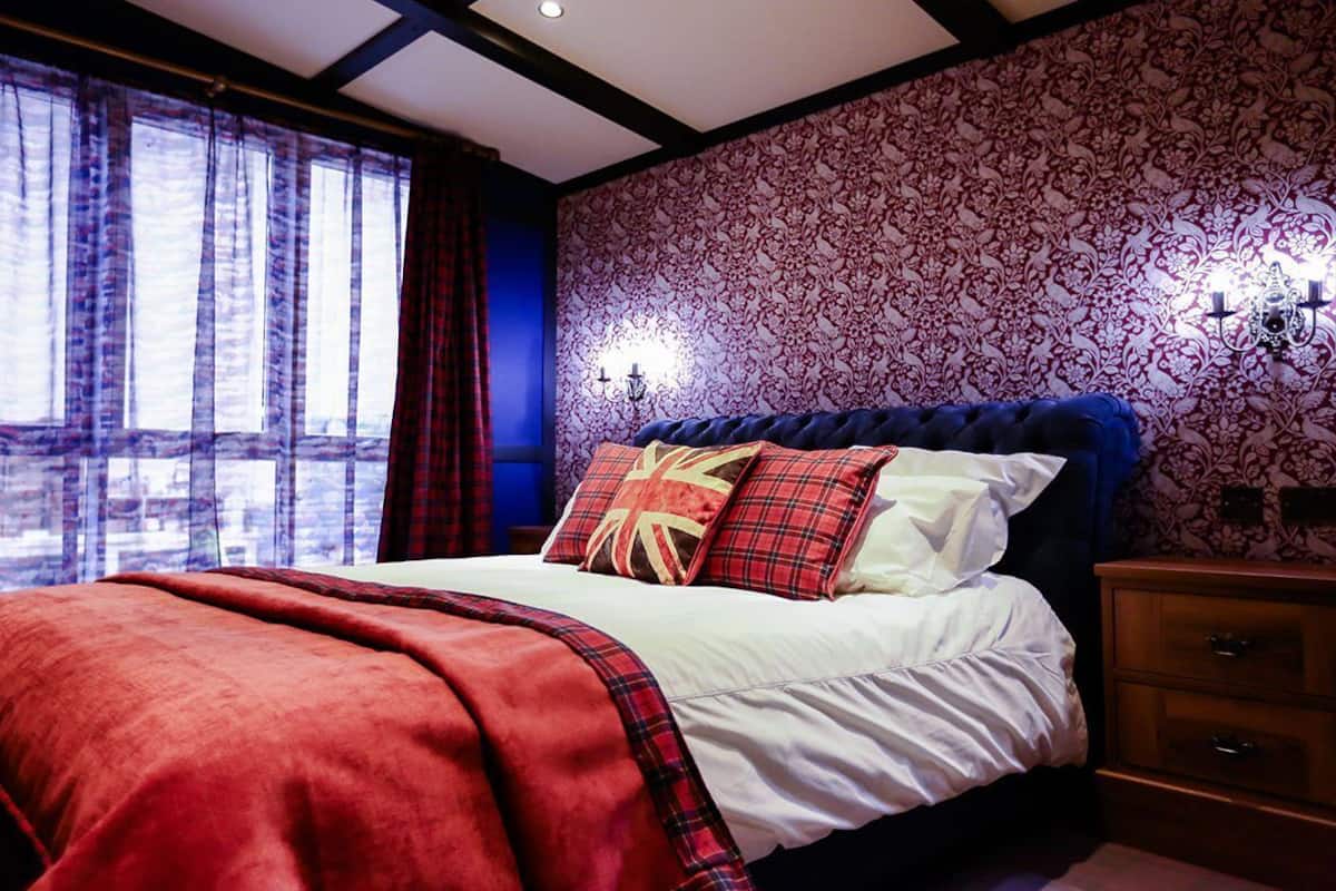 Un dormitorio con una cama grande, una ventana con cortinas, un papel tapiz estampado y una mesita de noche con una lámpara.