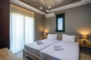 A hotel room with two beds, a chandelier, an air conditioner, a window with curtains, and bedside lamps.