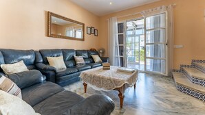 A living room with a dark leather sofa, a small table with a lace tablecloth, and a large window with curtains. - Holiday Home 'Townhouse beachside Fuengirola' with Shared Pool & Wi-Fi (Fuengirola)