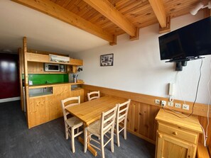 A compact kitchen with wooden cabinets, a microwave, and a green countertop. There is a dining table with four chairs and a television mounted on the wall.
