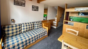 Quarto aconchegante com sofá xadrez, escada de madeira e cozinha compacta com detalhes verdes.