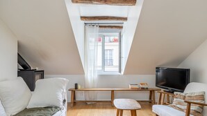 Une pièce mansardée chaleureuse avec un plafond en bois, une télévision, un canapé, une petite table et une fenêtre donnant sur les bâtiments environnants.