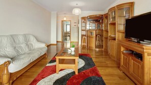 A living room with a sofa, a coffee table, a TV, wooden cabinets, and a kitchen area. - Apartment Costa Avila 2 with Sea View, Balconies & Wi-Fi (Caniço)