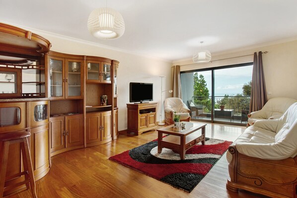 A living room with wooden cabinets, a flat-screen TV, a white sofa, a coffee table, and a large window with a view of greenery.