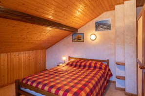 A bedroom with a wooden ceiling, a bed with a checkered blanket, a nightstand, a wall-mounted light, and a framed picture.