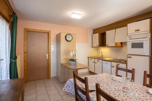 A kitchen with a clock, wooden cabinets, a dining table with a floral tablecloth, and a small potted plant.