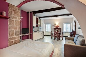 A bedroom with a bed, a TV, a stone wall, a kitchen, a dining area, and a living room with a sofa.