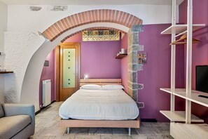 Un dormitorio con una cama, cabecera de madera, arco de ladrillo, pared morada, radiador blanco y una puerta de madera, además de un televisor sobre un mueble blanco.