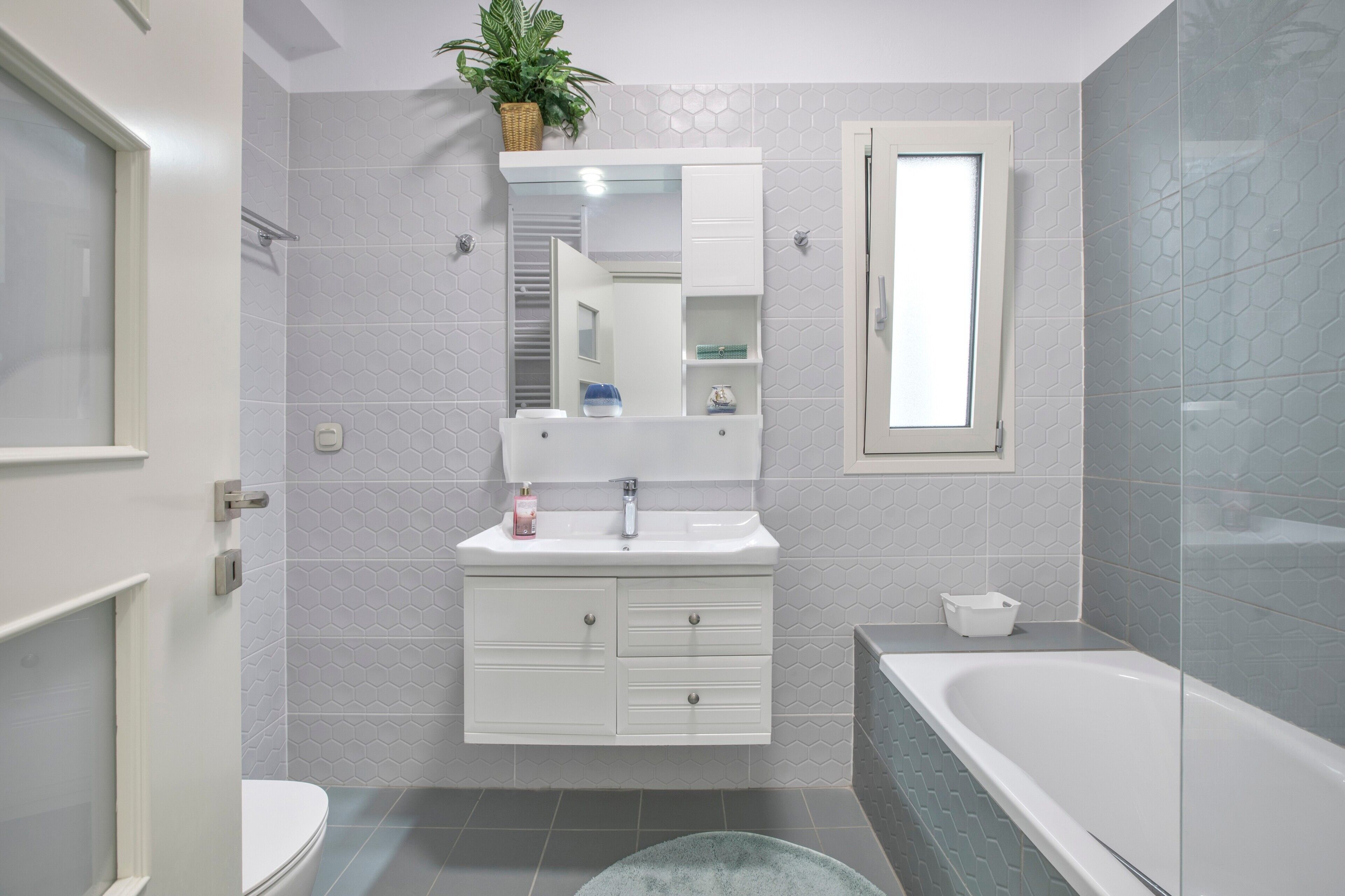 Et moderne badeværelse med et hvidt toiletbord, et stort spejl, en indbygget hylde, et badekar og en potteplante.